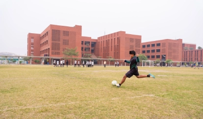 A student kicks a football on a grassy field near school buildings.