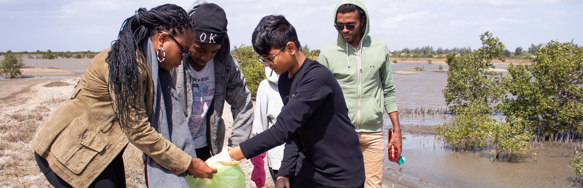 Students and teachers stand together outside near a lake, with the students collecting trash in a bag.