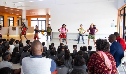 A group of young students dressed in traditional outfits stand on stage facing each other with a football between them, performing a drama in front of a wooden-panel backdrop.