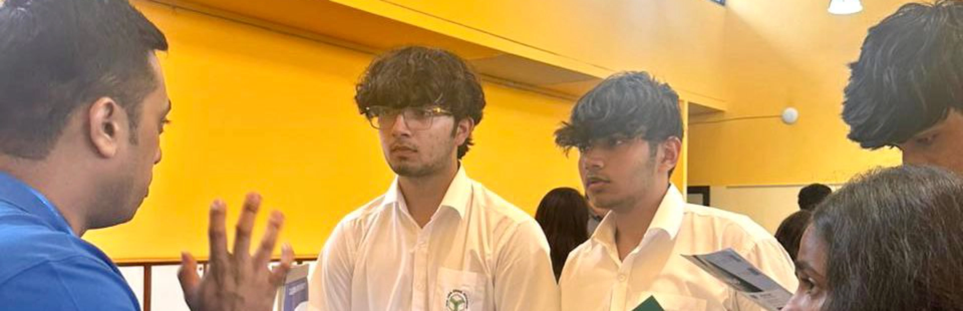 A group of students sitting in chairs with their backs to the camera. A woman is standing at the front of the room in front of a projection on the wall, addressing the students.