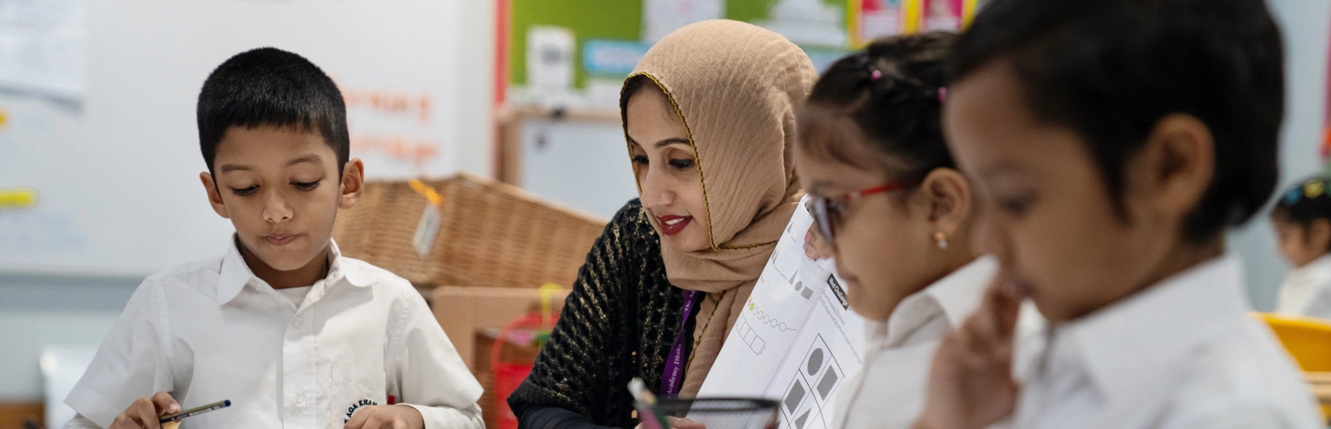 A teacher in a headscarf interacts with a group of children seated around a table, reviewing worksheets and helping them complete exercises in a classroom filled with educational displays.