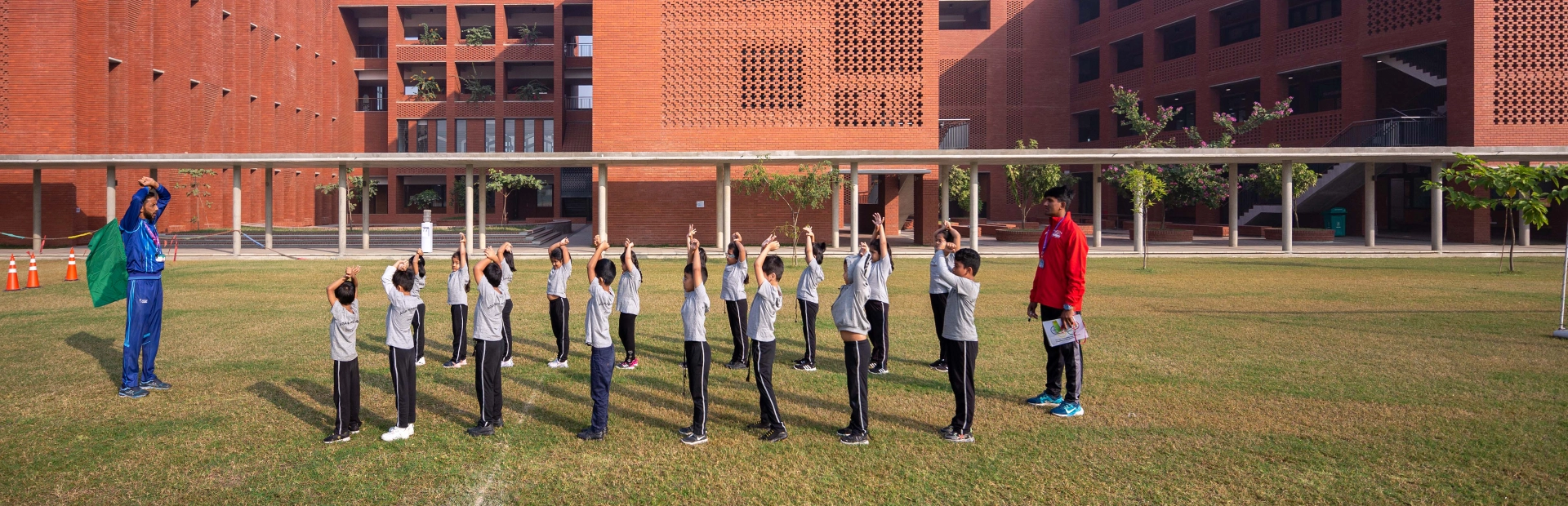 Junior School students standing in neat rows on a grassy field, following a warm-up exercise led by two instructors.
