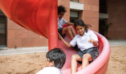 A group of young students dressed in traditional outfits stand on stage facing each other with a football between them, performing a drama in front of a wooden-panel backdrop.