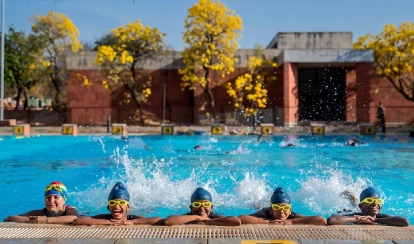 Two students stand together in the library reading a book, surrounded by bookshelves and other students browsing in the background.