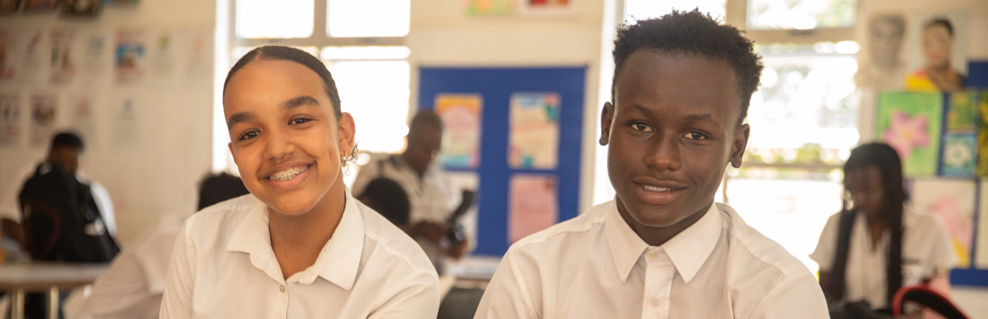 SlA student wearing glasses and a black headscarf holds up her colourful drawing featuring themes of nature, while another student works on art in the background.ider