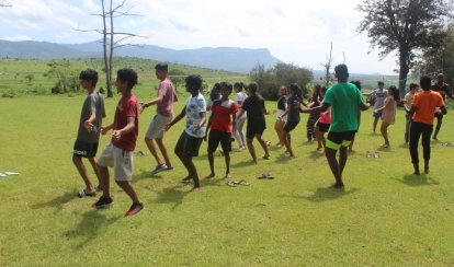 A student smiles while jumping over small yellow hurdles on the school field, with orange cones and buildings in the background.