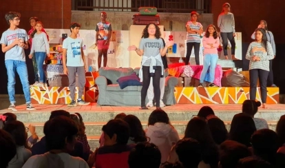 Two students stand together in the library reading a book, surrounded by bookshelves and other students browsing in the background.