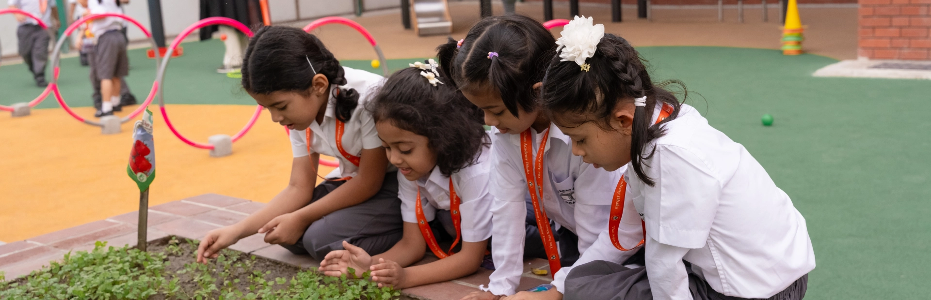 Four young students in school uniforms lean over a garden bed, closely observing and tending to small plants during an outdoor activity.