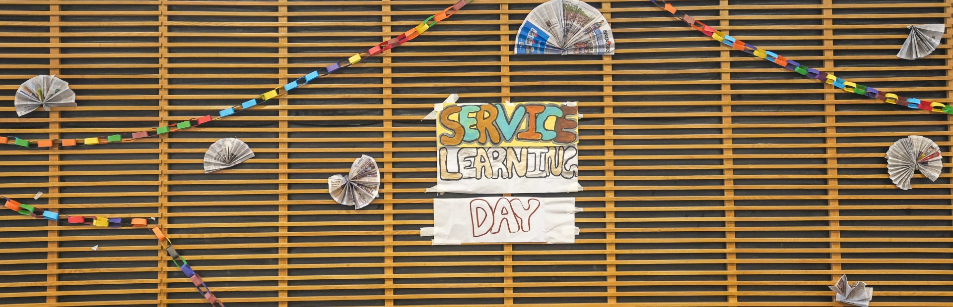 A decorative wall display with colorful paper chains and folded paper elements surrounding a hand-lettered sign that reads “Service Learning Day”.