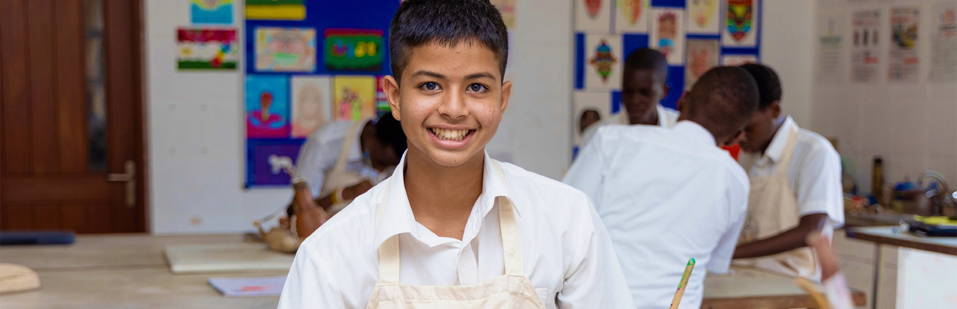 A young girl in a school uniform reads from an open book while her teacher attentively listens, resting her arms on the table and smiling in encouragement during a classroom reading session.