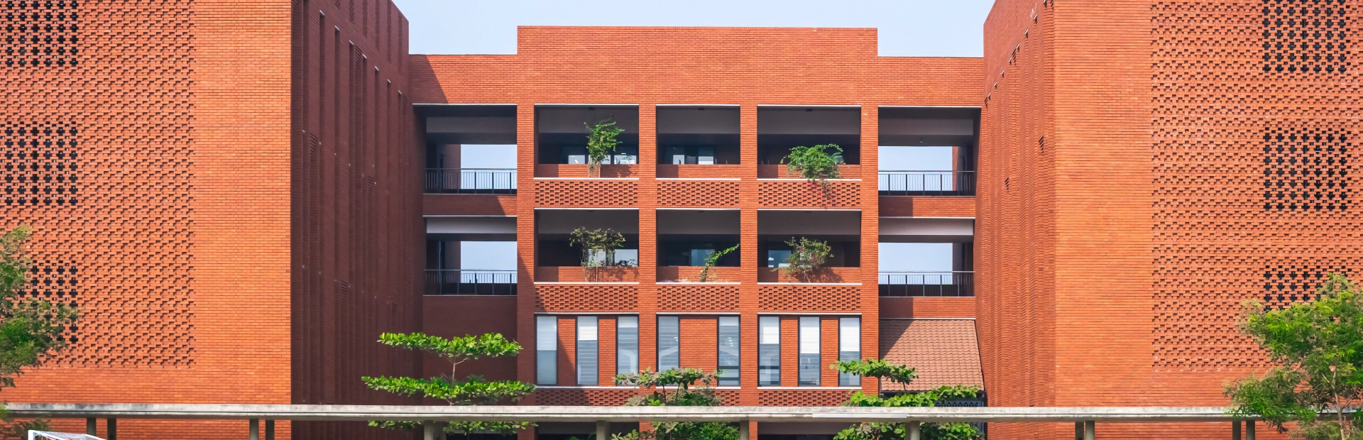 A wide view of the Aga Khan Academy Dhaka campus featuring its red-brick buildings, open green football fields, and clear blue sky.