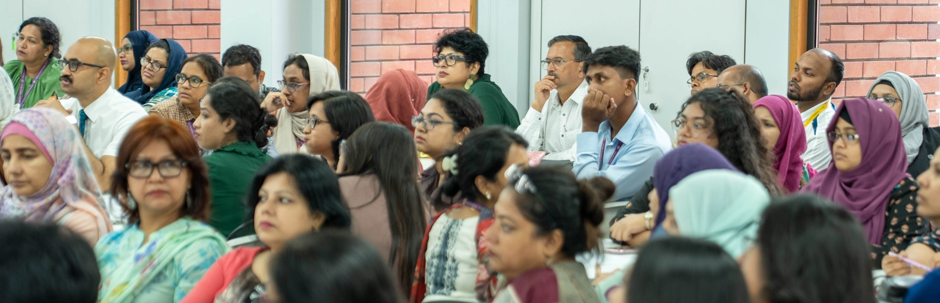A diverse group of teachers sits closely together in a bright meeting room, listening attentively to a presentation. The participants appear engaged, reflecting a collaborative professional development atmosphere.