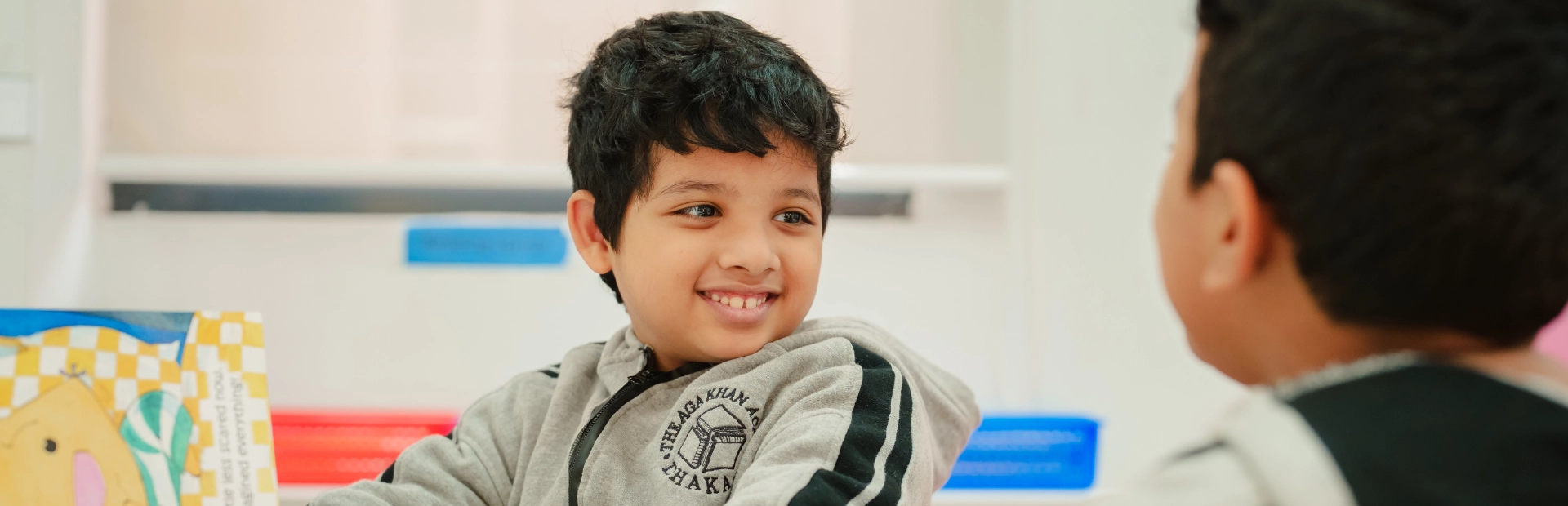 Two young students wearing grey school hoodies sit at a classroom table, smiling and discussing a colourful picture book together.