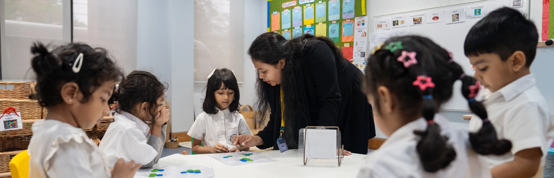 A teacher leans over a table to guide a group of young children during a classroom activity. The students, seated around the table, work with coloured counters on worksheets while the teacher offers individual support in a bright, welcoming learning environment.