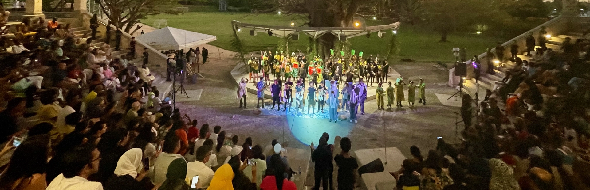 Three students sit at a table with laptops and notebooks in the library, focused on a group study session surrounded by bookshelves.