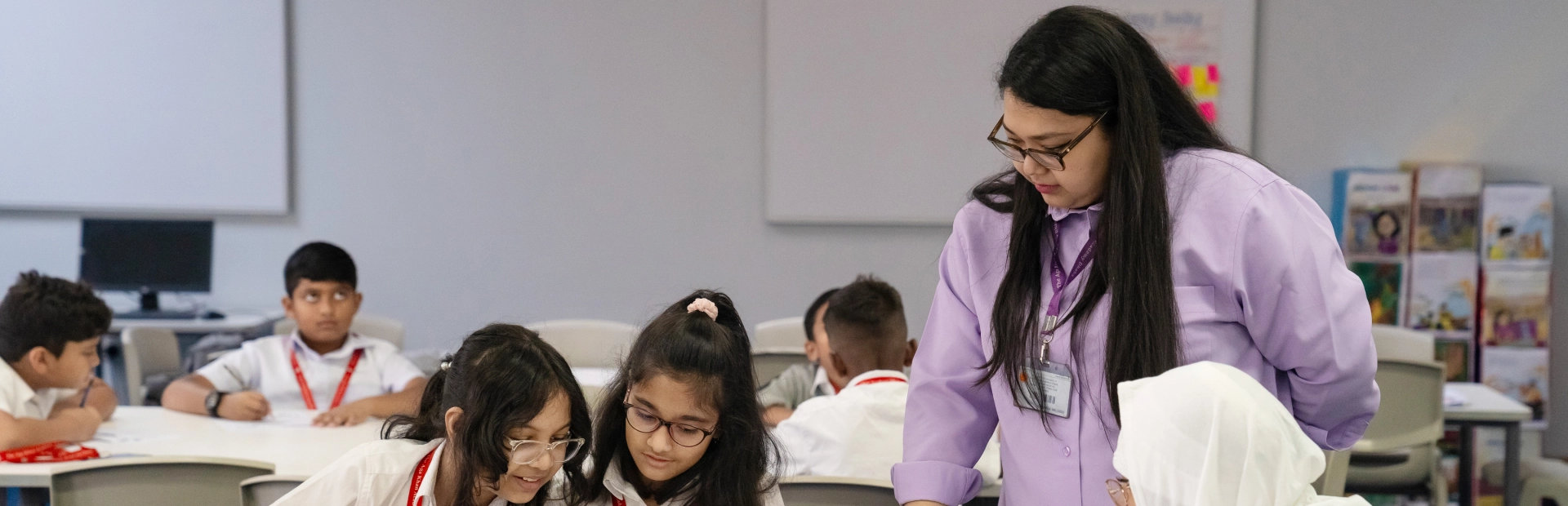 A teacher leans over a classroom table to assist a PYP student with a painting activity. The student attentively observes as the teacher demonstrates a technique with a paintbrush.