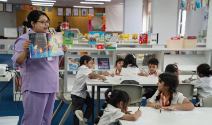 A teacher in a purple uniform holds up a colourful storybook while a group of young students sit at tables, listening and writing in the school library filled with books.