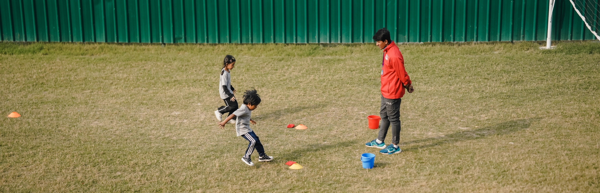 Two young students running between colored marker cones on a grassy field while a coach observes nearby.