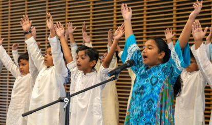 Junior School students stand on stage, raising their hands while singing into a microphone. One student in a bright blue patterned outfit leads the song, surrounded by classmates dressed in white traditional attire.