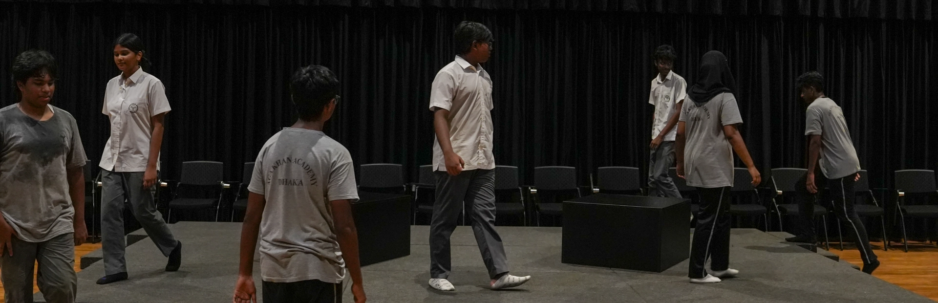 A group of students rehearsing on a stage with black curtains, walking and positioning themselves around simple stage blocks during a practice session.